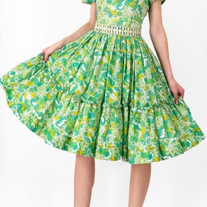 May include: A vintage-style green dress with a floral pattern. The dress features a square neckline, short sleeves, and a tiered skirt. A decorative belt cinches the waist. The model is wearing black strappy heels with red accents.