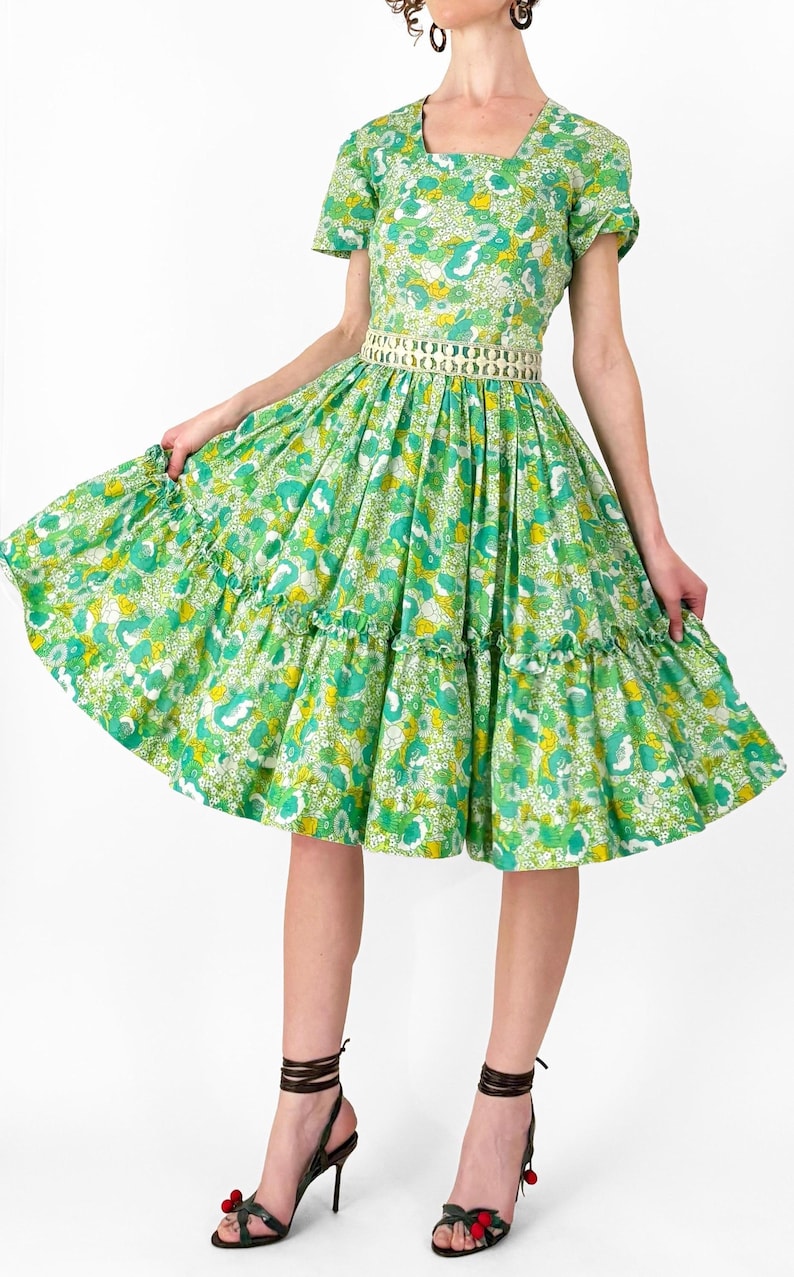 May include: A vintage-style green dress with a floral pattern. The dress features a square neckline, short sleeves, and a tiered skirt. A decorative belt cinches the waist. The model is wearing black strappy heels with red accents.
