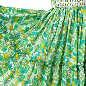 May include: A flowing green dress with a floral pattern in shades of green, yellow, and white. The dress features short sleeves, a decorative waist detail, and ruffled tiers. The fabric appears lightweight and airy, perfect for warm weather.