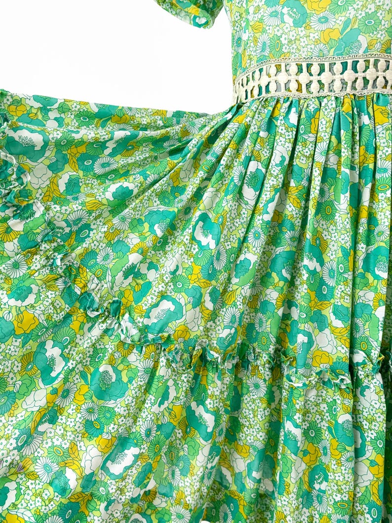 May include: A flowing green dress with a floral pattern in shades of green, yellow, and white. The dress features short sleeves, a decorative waist detail, and ruffled tiers. The fabric appears lightweight and airy, perfect for warm weather.