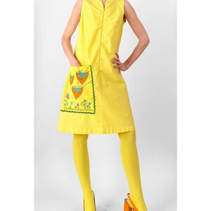 May include: A sleeveless yellow shift dress featuring a front zipper closure and a large patch pocket on the right side. The pocket is decorated with embroidered orange heart-shaped motifs and small blue and purple flowers with green stems.