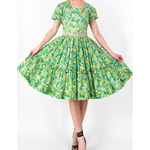 May include: A vintage green dress with a floral pattern, featuring a square neckline and short sleeves. The dress has a fitted bodice and a full skirt with ruffles. A decorative belt cinches the waist. The model is wearing black strappy heels.