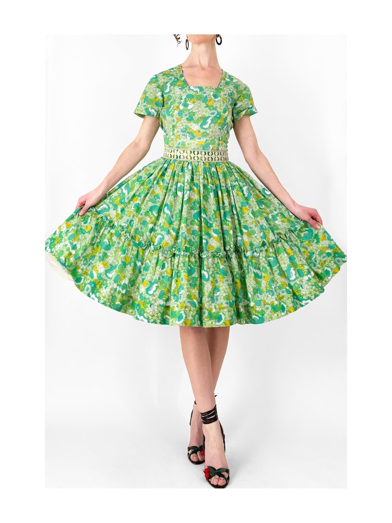 May include: A vintage green dress with a floral pattern, featuring a square neckline and short sleeves. The dress has a fitted bodice and a full skirt with ruffles. A decorative belt cinches the waist. The model is wearing black strappy heels.
