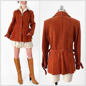 May include: A rust-colored suede jacket with a collared neckline, button closure, and a belt at the waist. The jacket has long sleeves with tie details at the cuffs. The jacket is displayed on a mannequin and a person.