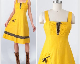 May include: A yellow sleeveless dress with a lace-up bodice, a small pocket with a bow, and a black trim around the hem.