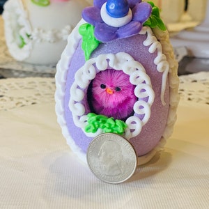 May include: A purple Easter egg with white lace trim and a pink fuzzy chick inside. The egg is decorated with a green leaf and a purple flower with a blue center. A US quarter is included for scale.