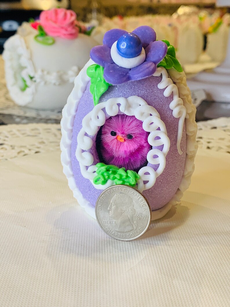May include: A purple Easter egg with white lace trim and a pink fuzzy chick inside. The egg is decorated with a green leaf and a purple flower with a blue center. A US quarter is included for scale.