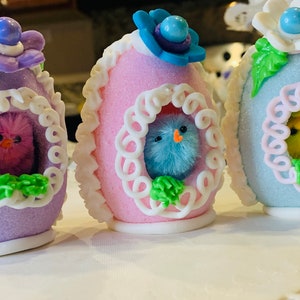 May include: Three decorated Easter eggs with chicks inside. The eggs are purple, pink, and blue with white frosting and green accents. Each egg has a different colored chick inside.
