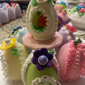 May include: A collection of decorated Easter eggs, some with glitter, some with frosting, and some with a bird design. The eggs are arranged on a white surface with a pink base.