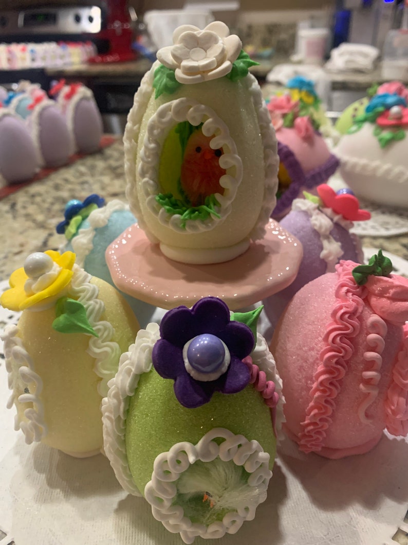 May include: A collection of decorated Easter eggs, some with glitter, some with frosting, and some with a bird design. The eggs are arranged on a white surface with a pink base.
