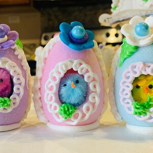May include: Three decorated Easter eggs with pastel colors and white trim. Each egg has a small fuzzy chick peeking out of a window. The chicks are pink, blue, and yellow.