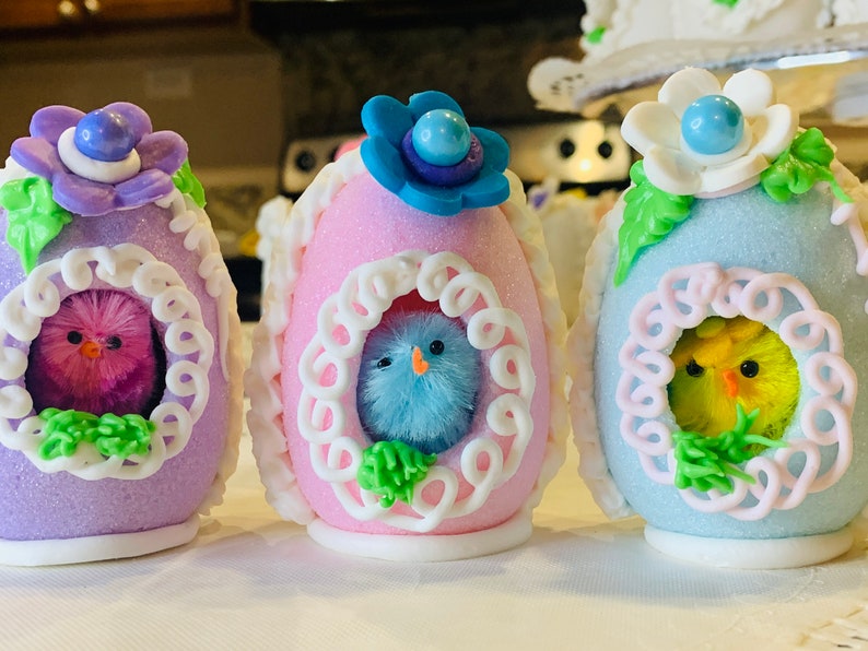 May include: Three decorated Easter eggs with pastel colors and white trim. Each egg has a small fuzzy chick peeking out of a window. The chicks are pink, blue, and yellow.