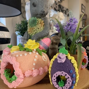 May include: Three decorated Easter eggs with a pink, purple, and yellow color scheme. The eggs are decorated with frosting, sprinkles, and flowers. The eggs are sitting on a wooden surface.