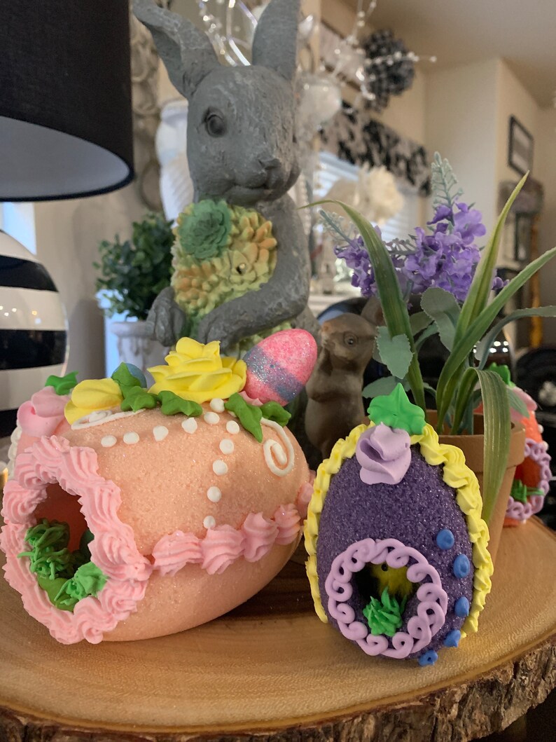 May include: Three decorated Easter eggs with a pink, purple, and yellow color scheme. The eggs are decorated with frosting, sprinkles, and flowers. The eggs are sitting on a wooden surface.