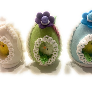Könnte beinhalten: Drei dekorative Ostereier in Pastellfarben. Jedes Ei zeigt ein kleines, flauschiges gelbes Küken, das durch eine dekorative Öffnung sichtbar ist. Die Eier sind weiß, grün und blau und mit floralen und spitzenartigen Details verziert, ideal für Ostern.