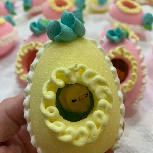 May include: A yellow Easter egg with a blue flower on top. The egg has a white border and a yellow swirl design. Inside the egg is a small yellow chick.