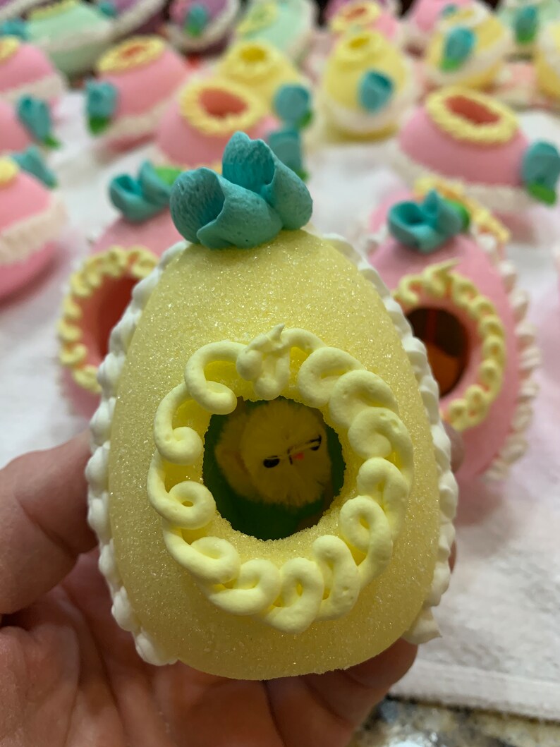 May include: A yellow Easter egg with a blue flower on top. The egg has a white border and a yellow swirl design. Inside the egg is a small yellow chick.