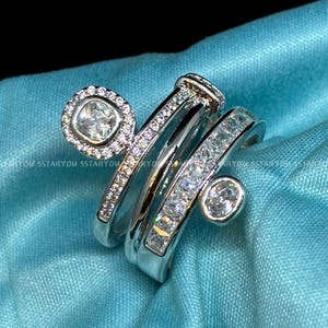 May include: A silver ring with three bands, each set with clear cubic zirconia stones. The top band features a larger, square-shaped stone.