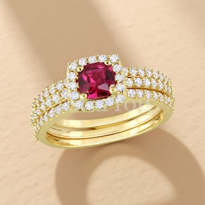 Könnte beinhalten: Ein goldfarbenes Ring-Set mit einem quadratischen, rubinroten Mittelstein, umgeben von einem Halo aus kleinen, klaren Edelsteinen. Das Band ist ebenfalls mit kleineren Edelsteinen besetzt. Das Set enthält ein passendes Band.