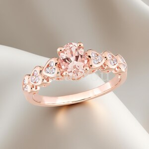 May include: Rose gold ring featuring an oval-cut, peach-colored gemstone as the centerpiece. The band is adorned with heart-shaped accents, each embellished with small, sparkling white stones. A delicate and romantic piece of jewelry.