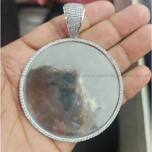 Puede incluir: Un colgante redondo de plata con una superficie lisa y vacía rodeada por un borde de pequeñas piedras transparentes.