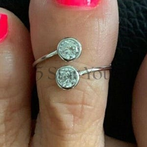 May include: A silver toe ring with two clear, round gemstones. The ring wraps around the toe, with one gem on top and the other below. The toe has bright pink nail polish.