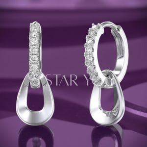 Puede incluir: Pendientes de aro plateados con un colgante en forma de lágrima. Los aros están engastados con pequeñas piedras brillantes. Los pendientes se muestran sobre un fondo morado. El texto "STAR Y" es visible.