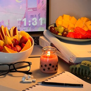 Op de afbeelding: Een witte kom gevuld met gesneden fruit, waaronder aardbeien, sinaasappels en appels. De kom staat op een tafel met een laptop, een kaars, een notitieboekje en een bril. Het laptopscherm toont een digitale klok die 1:31 uur 's ochtends aangeeft.