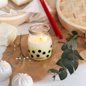 Op de afbeelding: Een gele kaars met zwarte stippen in een glazen pot met de tekst "ASIANBORAGIRL" op de bodem. De kaars is aangestoken en er zijn witte gestoomde broodjes en eetstokjes op de achtergrond.