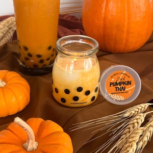 Op de afbeelding: Een oranje kaars in een glazen pot met zwarte stippen, naast een glas oranje drank met donkere boba-parels. Een kleine pompoen en een grotere pompoen staan ook in de afbeelding. Het kaarslabel luidt "Rich Pumpkin Thai".