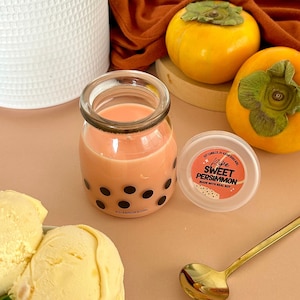 Op de afbeelding: Een glazen pot met een roze vloeistof en zwarte stippen, met het label "Asianbobagirl". Ernaast ligt een wit deksel met de tekst "Ripe Sweet Persimmon Made with Real Fruit". Op de achtergrond zijn twee oranje kaki's en een kom vanille-ijs te zien.