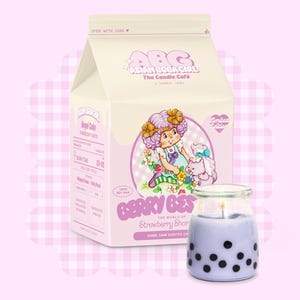 Op de afbeelding: Een paarse en witte melkpakvormige kaars met een cartoonillustratie van een meisje en een hond. De kaars is gelabeld met "Berry Best" en "Strawberry Shortcake" met de tekst "Angel Cake Scented Candle".