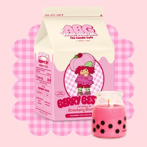 Op de afbeelding: Een roze en witte melkpak-kaars met de tekst "Asian Boba Girl The Candle Cafe" en een cartoon-aardbei-personage. De kaars is gevormd als een melkpak en heeft een frambozen-taartgeur.