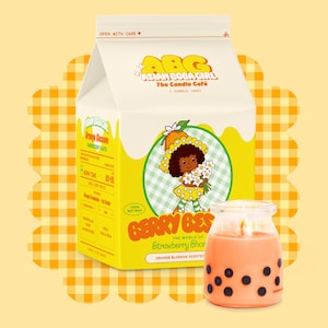 Op de afbeelding: Een kaars en een doos met kaarsen met de tekst "ASIAN BOGA GIRL The Candle Café". De kaars zit in een glazen pot met zwarte stippen. De doos is geel met een cartoonmeisje-illustratie.