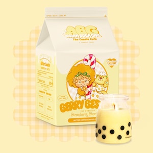 Op de afbeelding: Een gele en witte melkpakkerze met een cartoonfiguur op de voorkant. De kaars is gelabeld "Berry Best" en "Strawberry Shortcake" en is een boterkoekgeurkaars. De kaars zit in een glazen pot met zwarte boba-thee stippen.