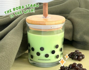 May include: A green and black candle with a boba straw wick. The candle is in a glass jar with a bamboo lid. The text "ASIANBOBAGIRL" is printed on the bottom of the jar. There are two piles of black boba pearls on a light green surface.
