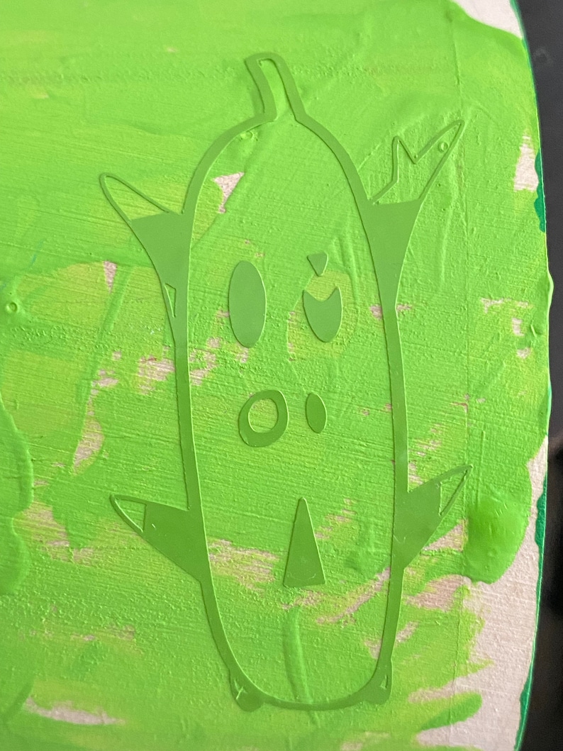 May include: Green painted wooden surface with a green silhouette of a cartoon character with a large head, two eyes, a nose, and a mouth. The character has two arms and two legs.