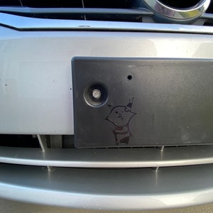 May include: A silver Nissan car with a black license plate. The license plate has a sticker of a cartoon cat with a black and white striped shirt.