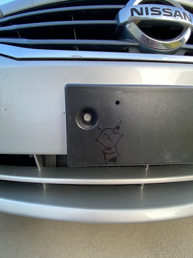 May include: A silver Nissan car with a black license plate. The license plate has a sticker of a cartoon cat with a black and white striped shirt.