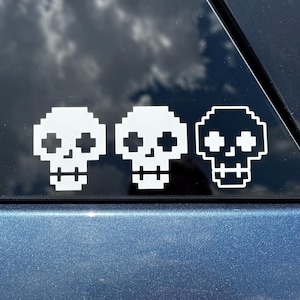 May include: A white vinyl decal featuring three pixelated skull designs arranged in a horizontal row. Each skull is rendered in a blocky, 8-bit style with cross-shaped eye sockets and a simple rectangular jaw line, applied to a dark glass surface.