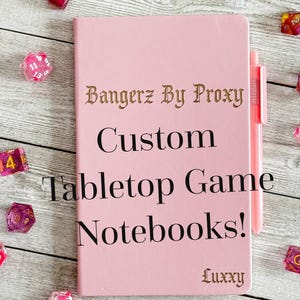 May include: A pink notebook with the text "Bangerz By Proxy Custom Tabletop Game Notebooks!" and the name "Luxxy" in gold lettering. A pink pen is attached to the notebook. Scattered around the notebook are several pink and purple dice.