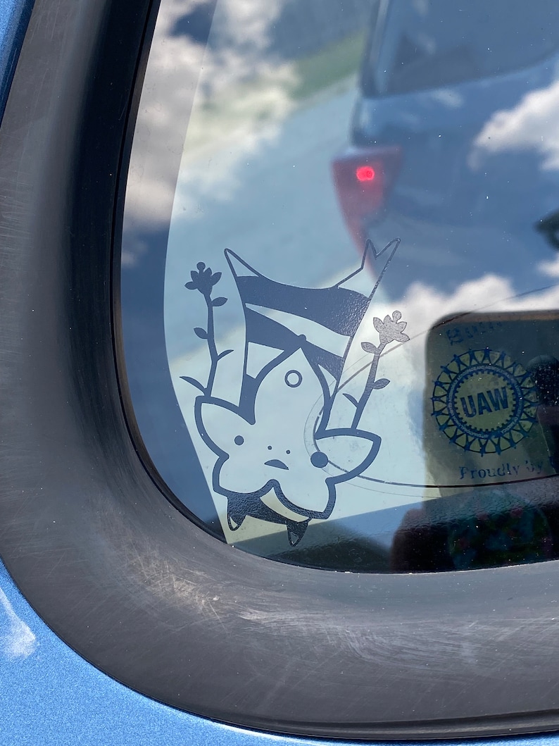 May include: A black and white sticker of a cartoon character with a star-shaped head and a striped body. The character is upside down and has two small flowers growing from its head. The sticker is on a car window with a UAW sticker in the background.