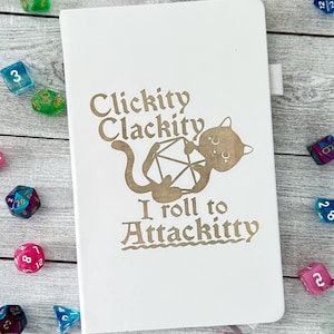 May include: A white notebook with a gold design of a cat holding a twenty-sided die. The text on the notebook reads "Clickity Clackity I roll to Attackitty".