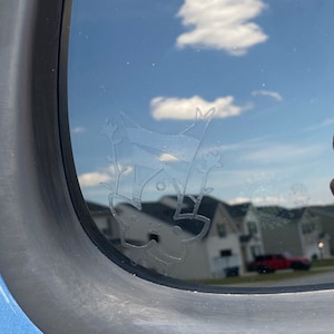 May include: A close-up of a car window with a white etched design of a cat hanging upside down. The cat has a round head, two ears, and a tail. The design is on the inside of the window, and the reflection of the sky and clouds can be seen through the glass.