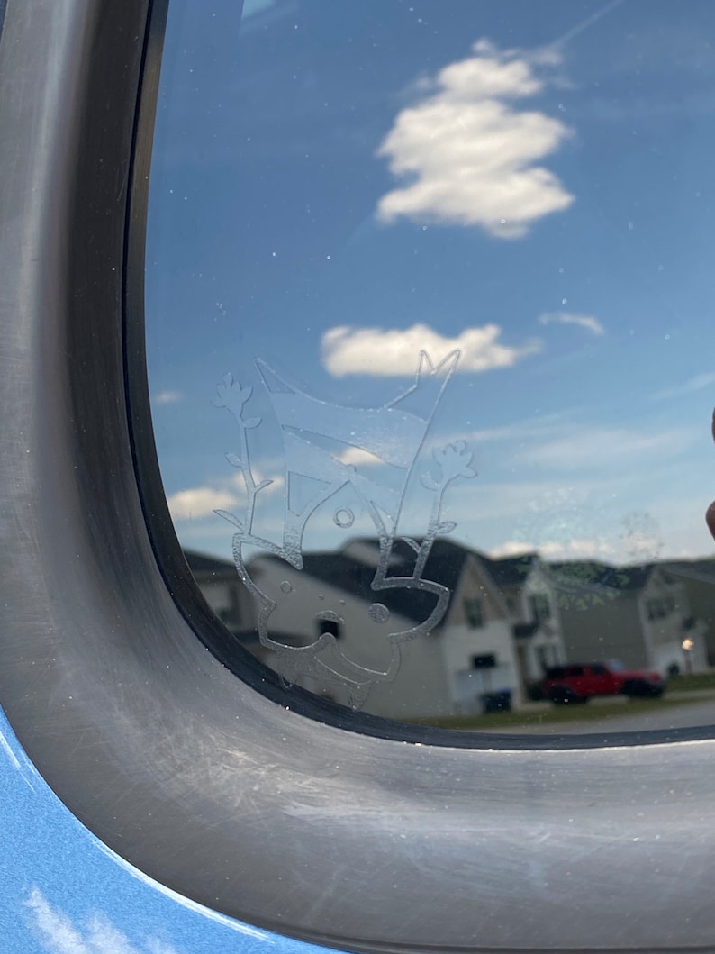 May include: A close-up of a car window with a white etched design of a cat hanging upside down. The cat has a round head, two ears, and a tail. The design is on the inside of the window, and the reflection of the sky and clouds can be seen through the glass.
