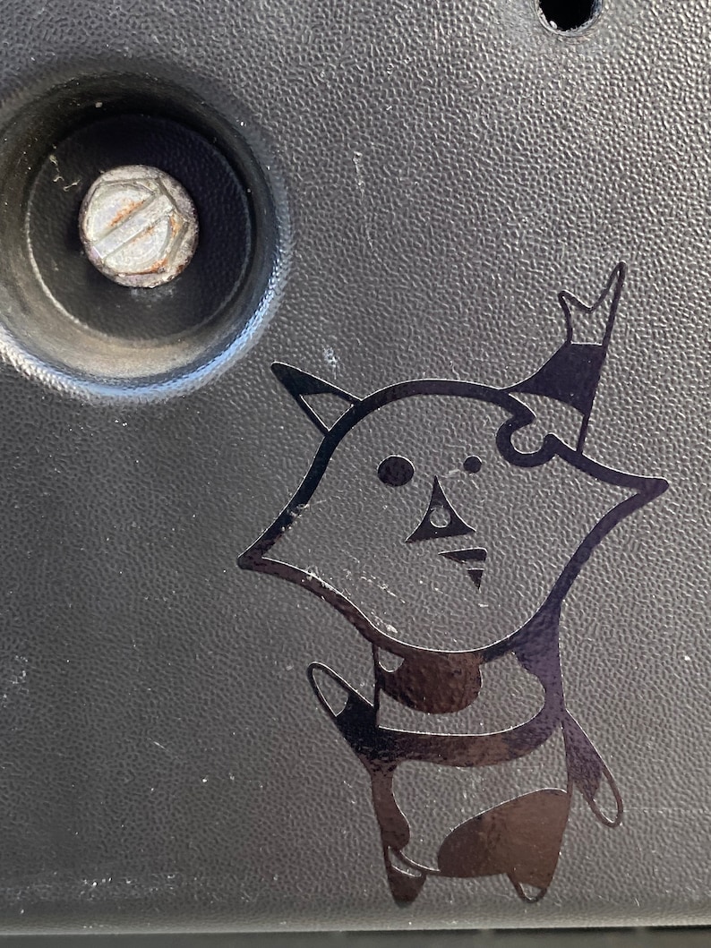 May include: Black and white sticker of a cartoon character with a crown on its head. The character is wearing a striped shirt and has a large, round head.