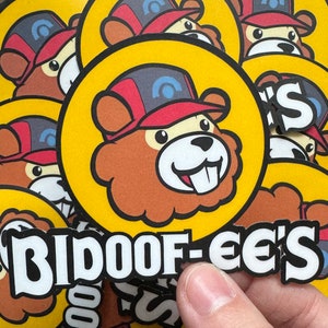 May include: A close-up of a sticker with the text "Bidoof-ee's" in white letters on a black background. The sticker is placed on a yellow background with a cartoon image of a brown bear with a red and white hat.