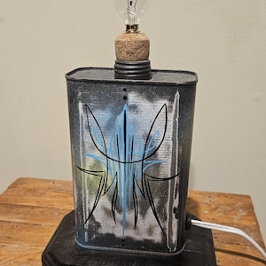 May include: A black and white lamp with a blue pinstripe design on a metal can base. The lamp has a clear light bulb and a cork top. The lamp is sitting on a wooden table.