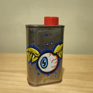 May include: A rectangular metal container with a red screw-top cap. The front surface features a hand-painted graphic of a white eyeball with a blue iris and yellow wings, surrounded by small blue dots against the metallic gray background.