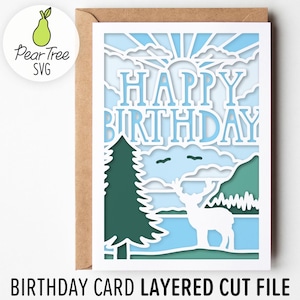 May include: A layered paper birthday card featuring a white die-cut design of a deer standing in a forest landscape with a sunburst and clouds in the background. The text HAPPY BIRTHDAY is cut out in the upper section. The card is set against a brown envelope.
